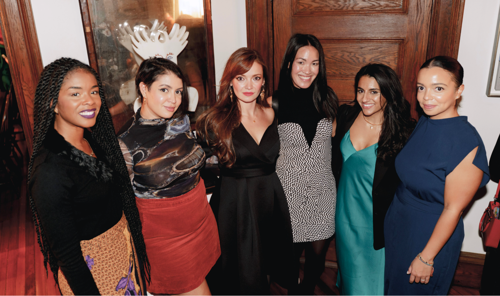 Group of six women posing together in a formal setting with wooden doors in the background.